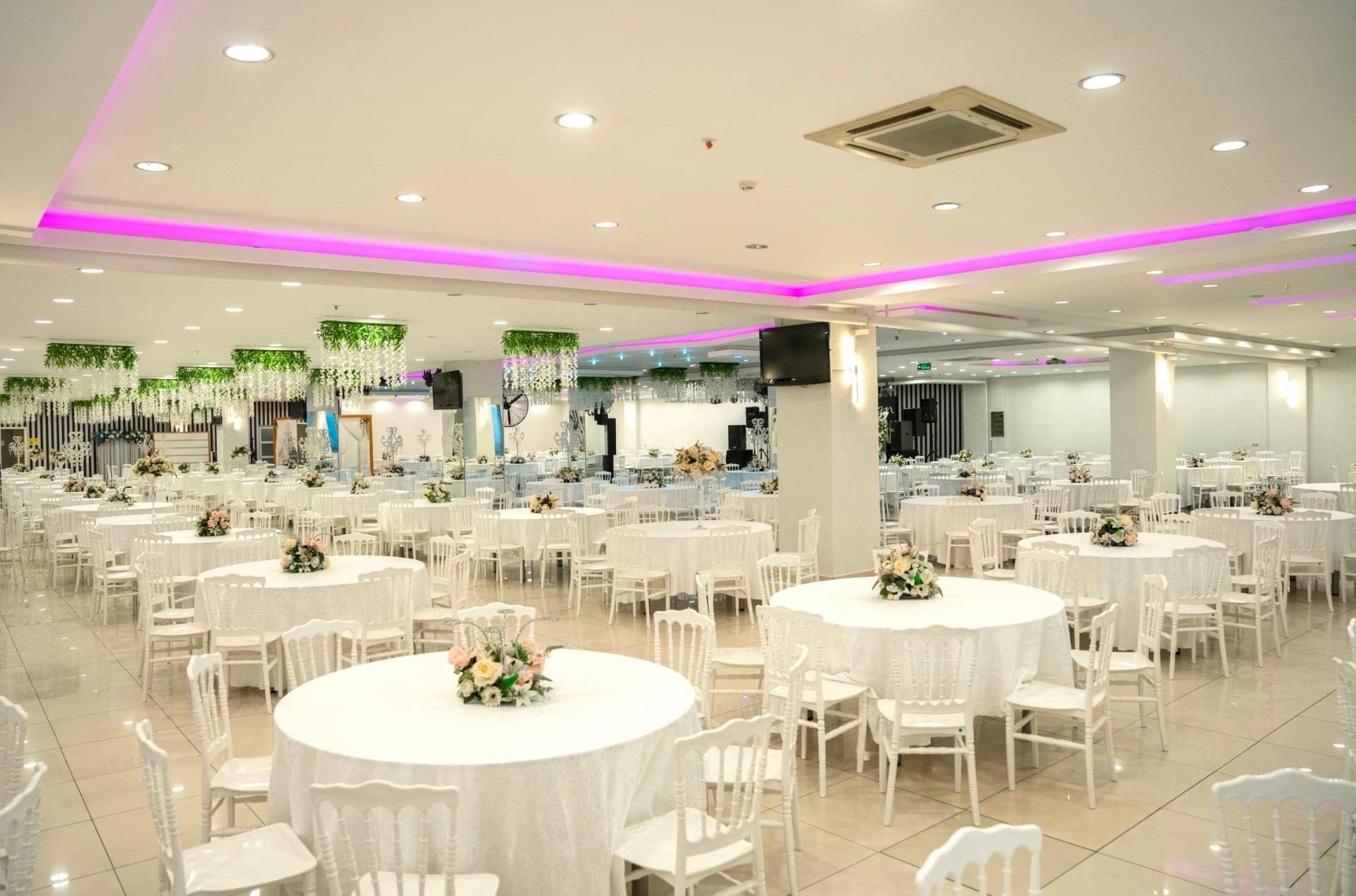 Pembe tavan ışıkları altında beyaz örtülü yuvarlak masalarla kurulmuş salon