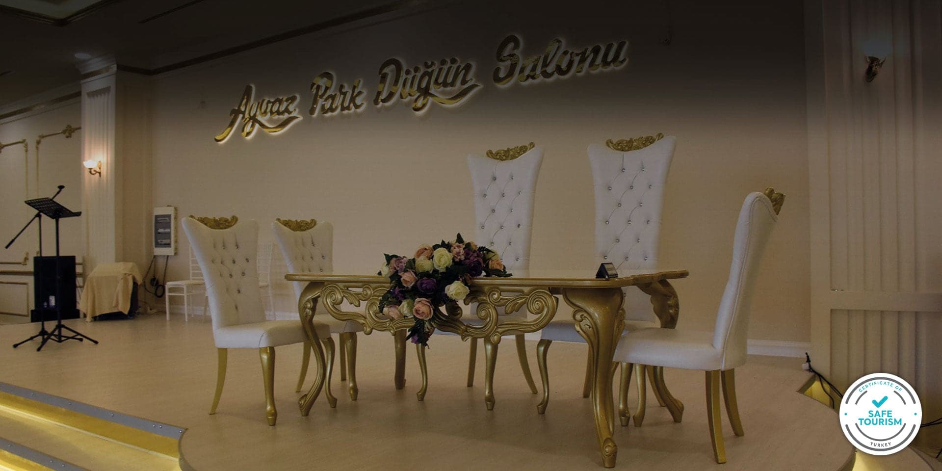 Düğün salonunda altın ayaklı gelin damat masası, beyaz koltuklar ve çiçek aranjmanı