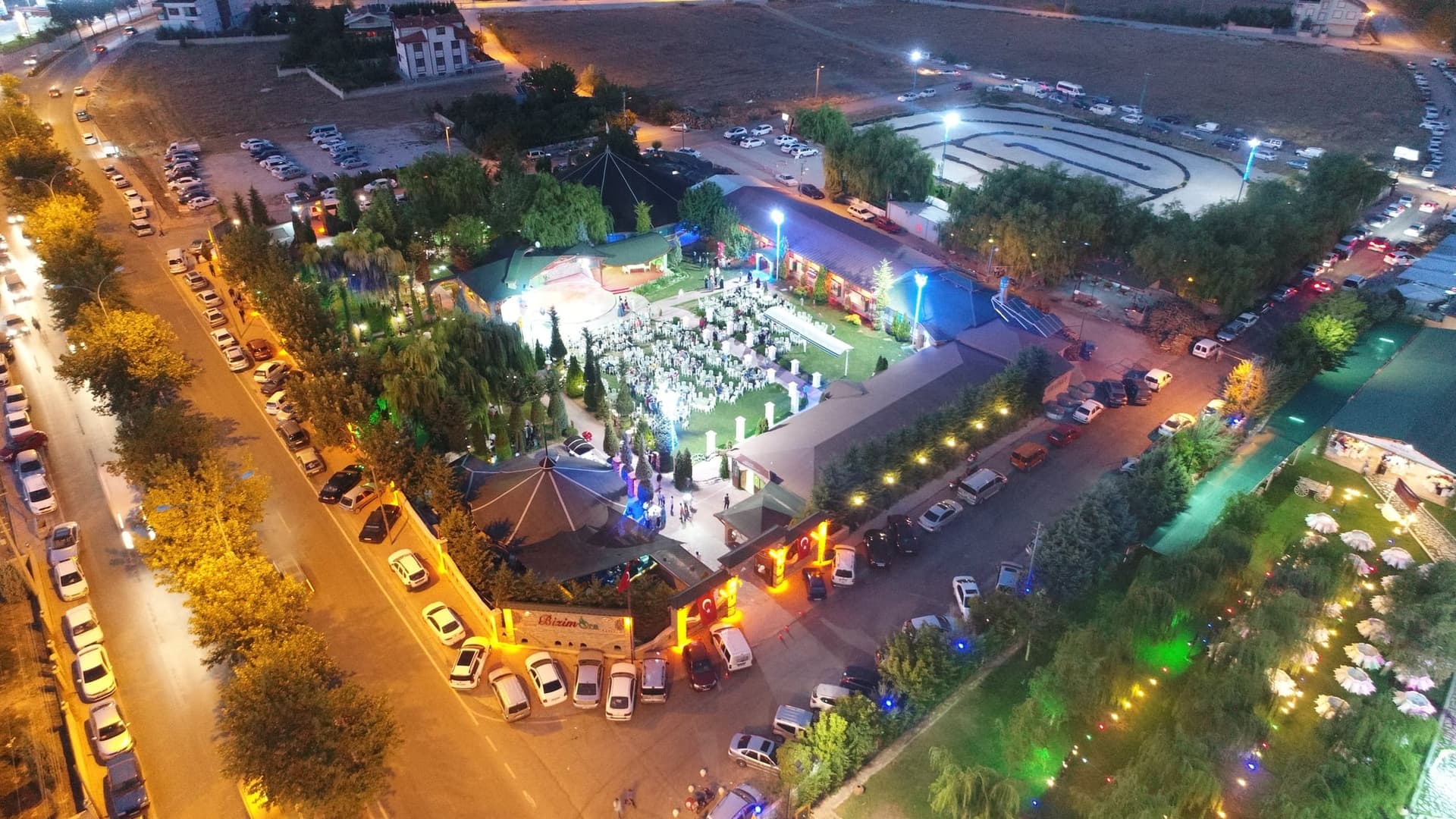 Gece havadan çekilmiş ışıklandırılmış düğün bahçesi, çimdeki masalar ve çevresindeki otopark