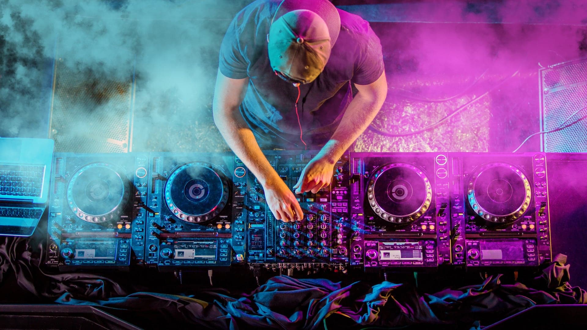 Dumanlı sahnede pembe ve mavi ışık altında dört çalarlı kurulumda çalan DJ, yukarıdan