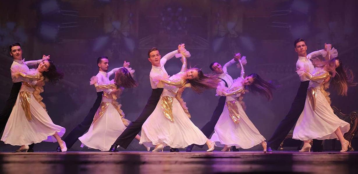 Mor ışıklı sahnede beyaz etekli kadınlar ve siyah giyimli erkeklerle eşli salon dansı gösterisi