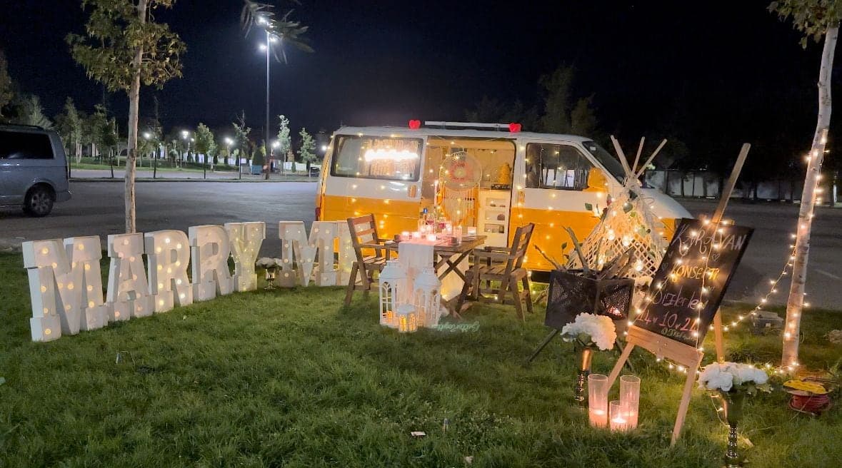Gece ışıklandırılmış sarı beyaz karavan, önünde iki kişilik masa ve ışıklı Marry Me harfleri