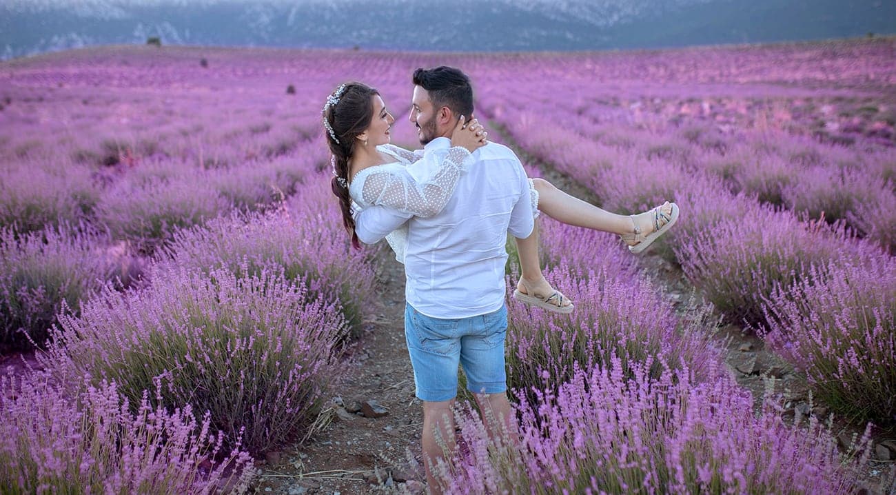 Lavanta tarlasında sıraların arasında gelini kucağında taşıyan damat, arkada dağlar