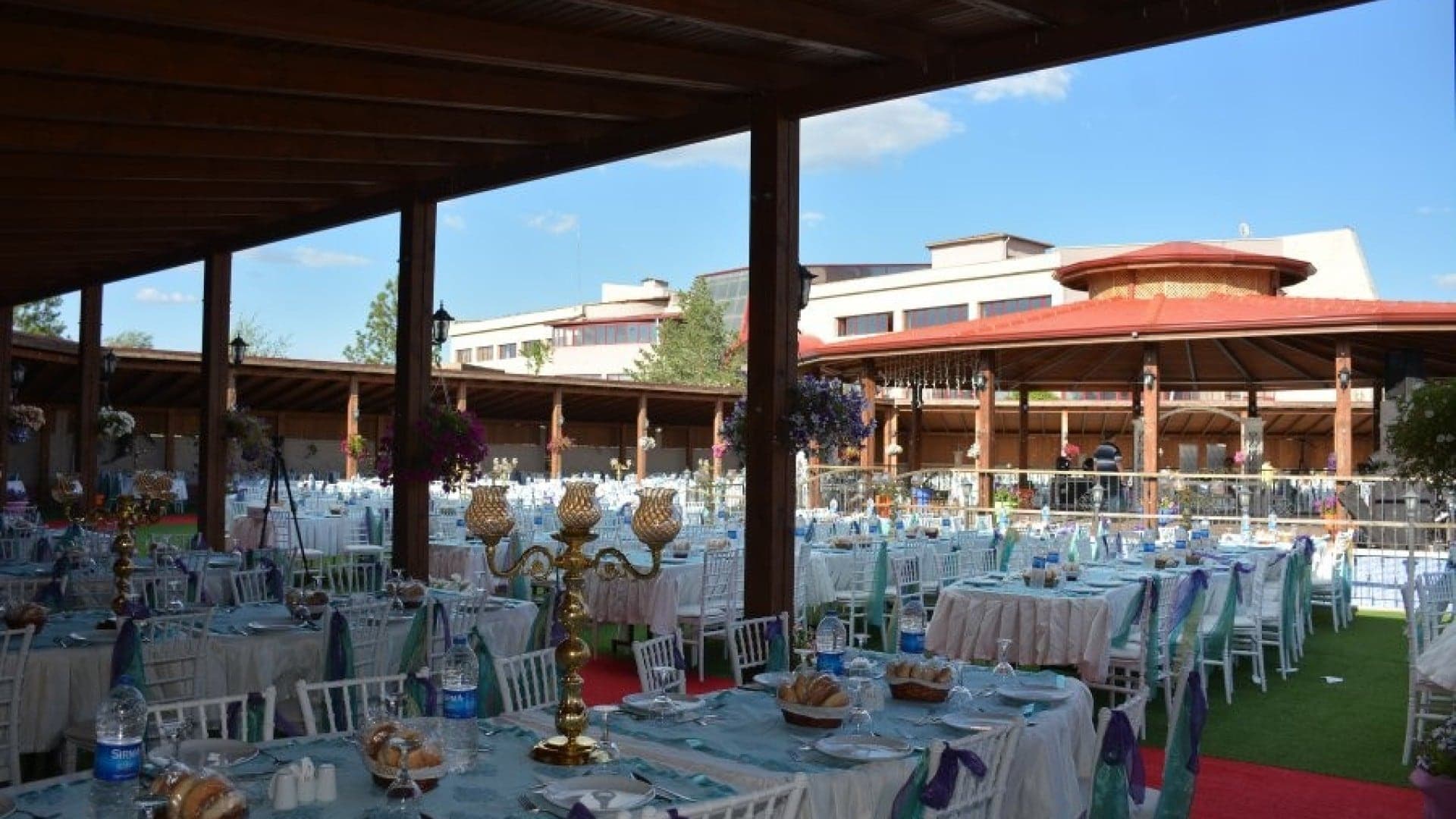 Ahşap veranda altında mavi örtülü uzun masalar ve şamdanlar, karşıda kırmızı çatılı köşk