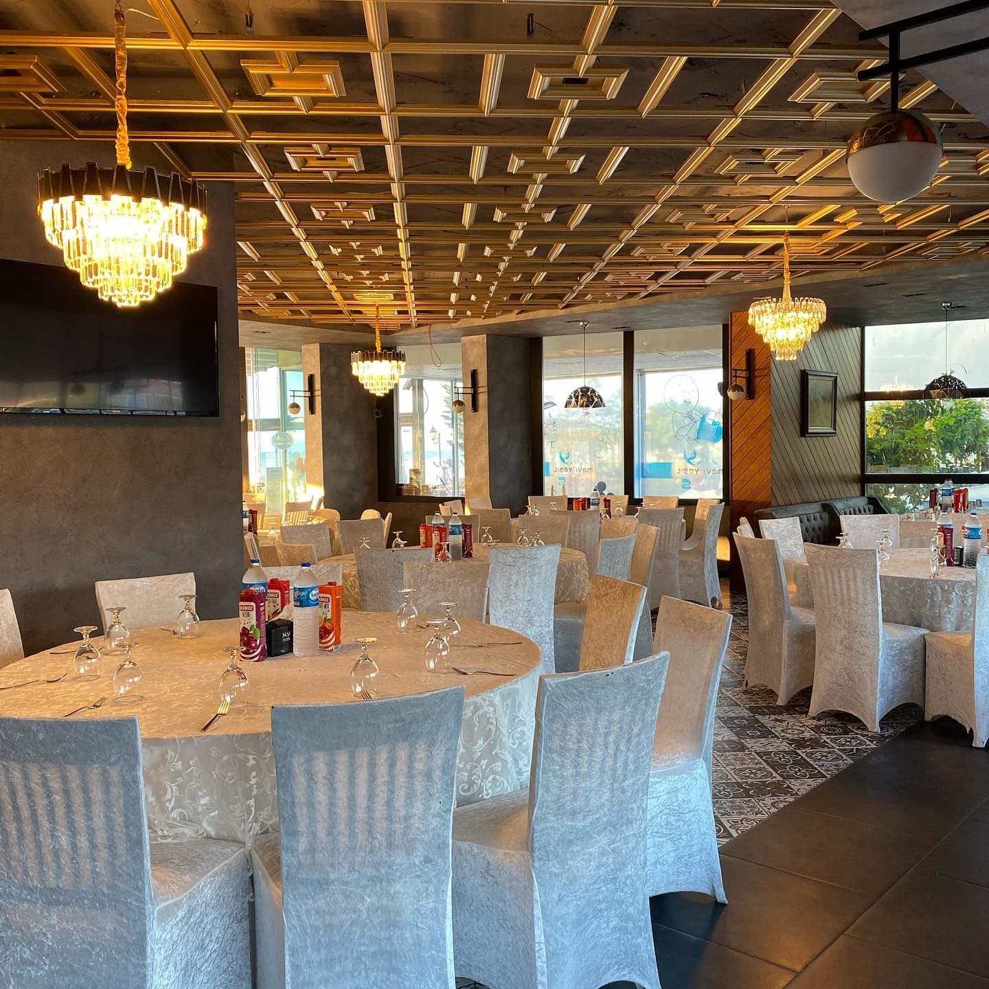 Beyaz örtülü sandalyeler ve yuvarlak masalarla kurulmuş restoran salonu, tavanda kristal avizeler